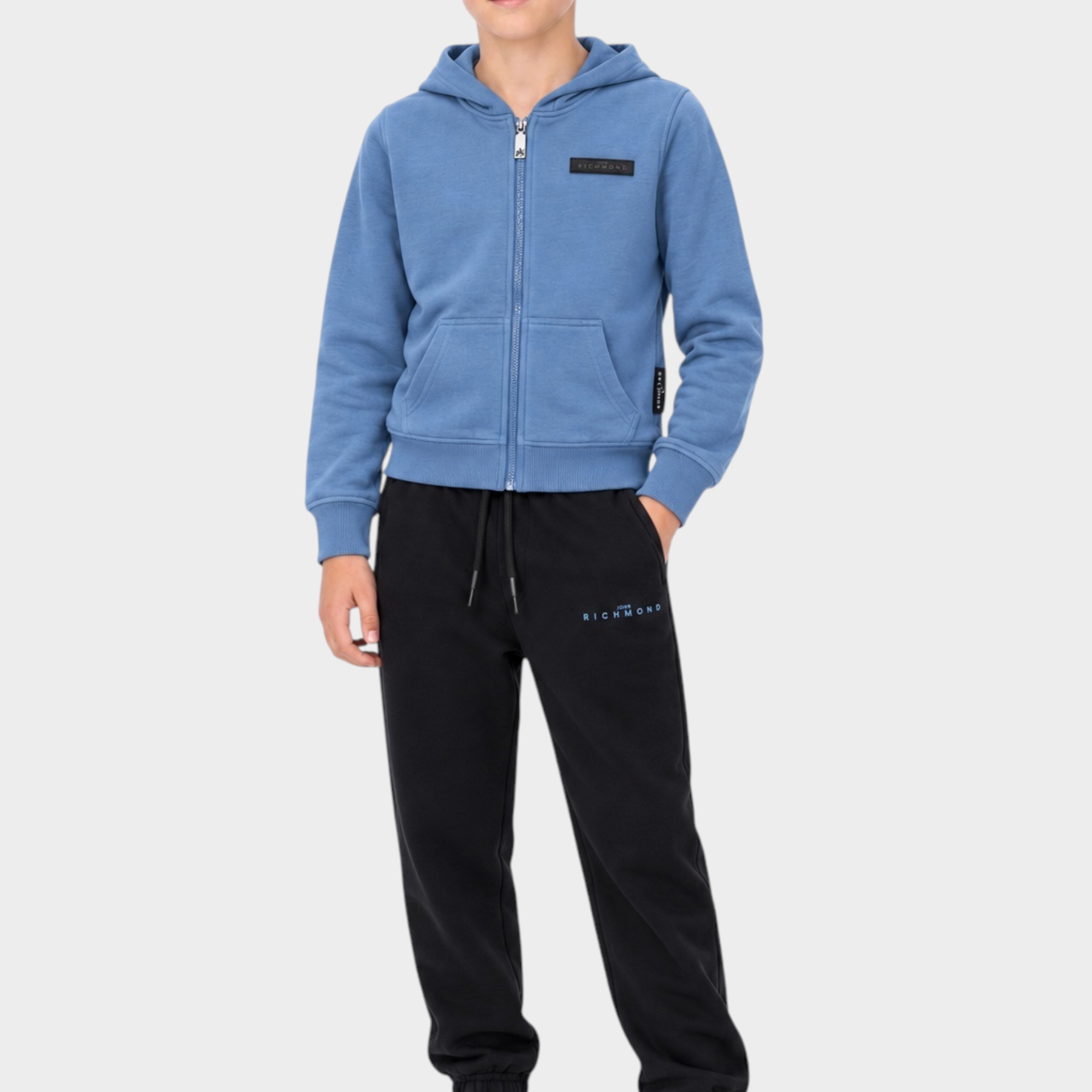 Ragazzo indossa completo sportivo Follower SRL con felpa azzurra con zip e pantaloni neri con logo RICHMOND