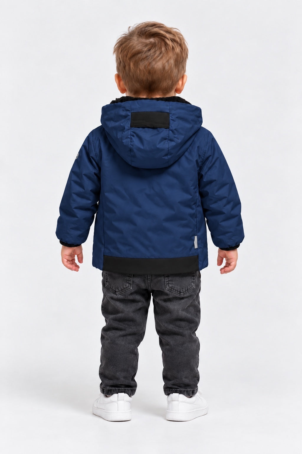 Bambino di spalle indossa un giubbino blu SUNS BOARDS in velour con cappuccio, jeans grigi e scarpe bianche