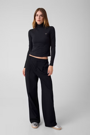 Modella indossa maglia lupetto nera in microfibra GUESS con logo metallico sul petto e pantaloni neri a gamba larga