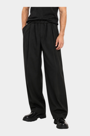 Modello indossa pantaloni baggy-fit con pinces Follower SRL in colore scuro con t-shirt nera e scarpe eleganti